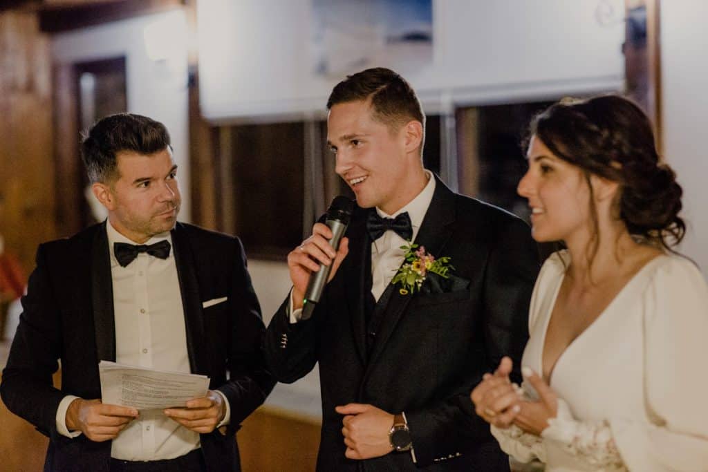 Top8 master of ceremonies in 2026 / the best masters of ceremonies for your wedding 12 Three elegantly dressed people are talking, one man is holding a microphone, another is holding papers, and a woman is listening.