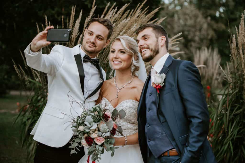 Top8 master of ceremonies in 2026 / the best masters of ceremonies for your wedding 11 Three people, including a bride, take a selfie in a green garden at a wedding.