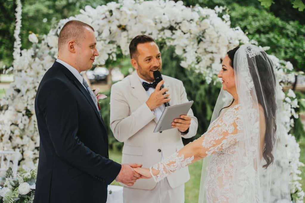 Top8 master of ceremonies in 2026 / the best masters of ceremonies for your wedding 6 Wedding ceremony where the couple exchanges vows under a floral arch, with a master of ceremonies.