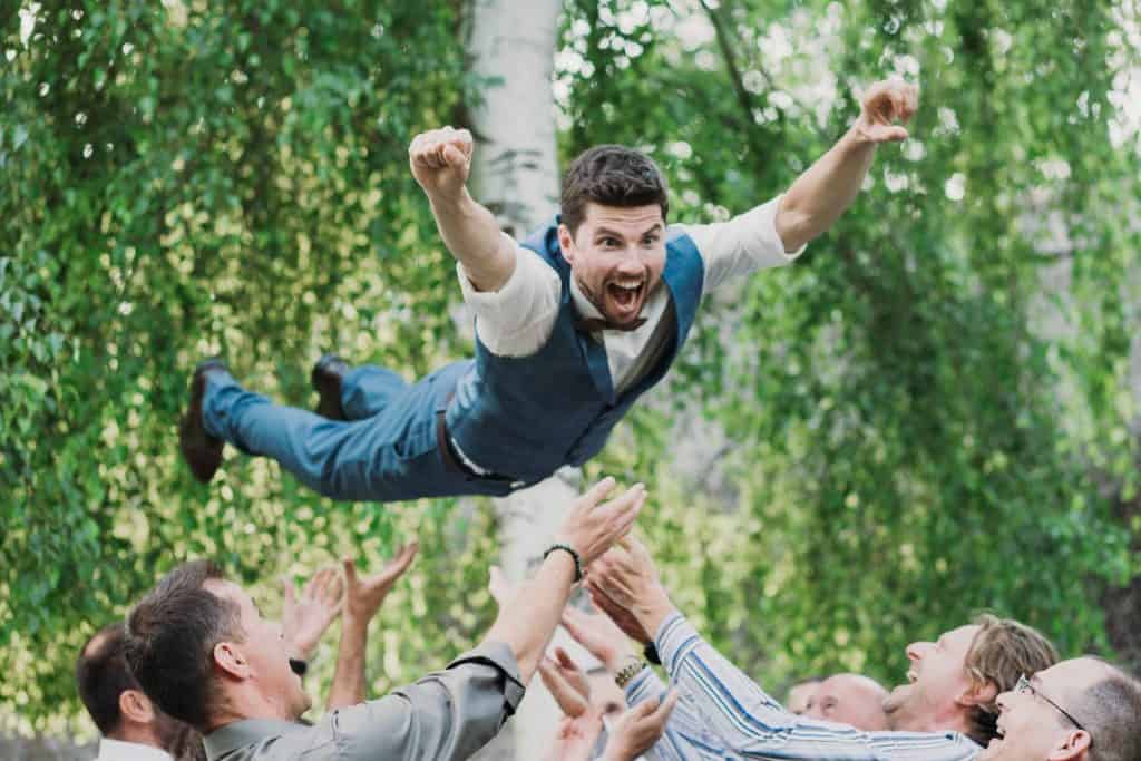 Top8 master of ceremonies in 2026 / the best masters of ceremonies for your wedding 15 Several people are throwing a man in a suit into the air next to green leaves.