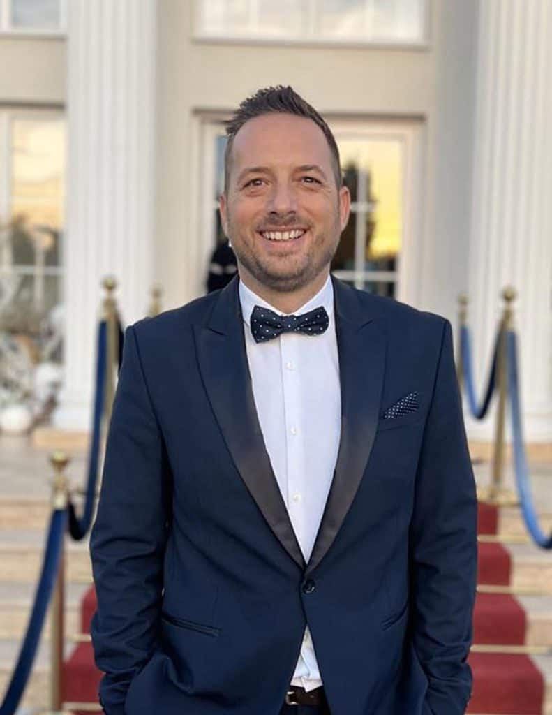 Top8 master of ceremonies in 2026 / the best masters of ceremonies for your wedding 4 Smiling man in a navy blue suit and polka dot bow tie in front of a stepped entrance.