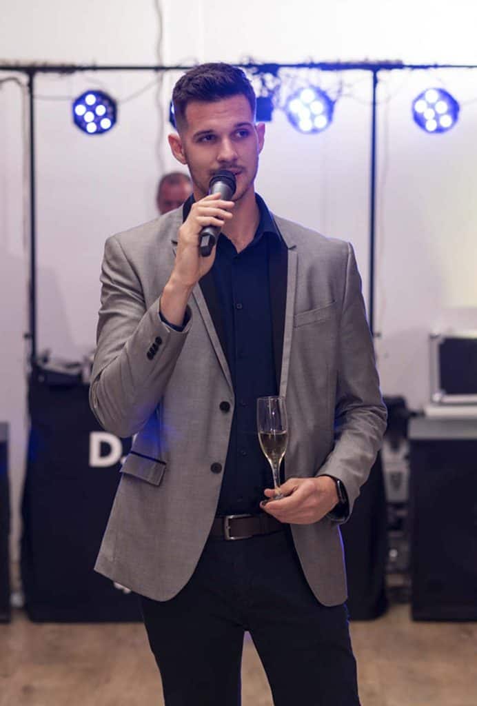 Top8 master of ceremonies in 2026 / the best masters of ceremonies for your wedding 16 Young man in a gray suit holding a microphone and a champagne glass at an indoor event.