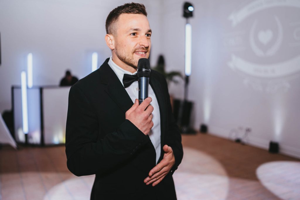 Férfi fekete öltönyben és masnival mikrofont tartva beszél rendezvényen, háttérben világítás és grafika.