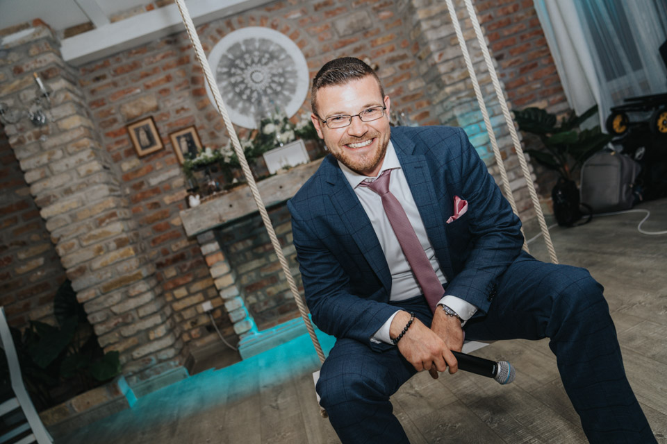 Férfi öltönyben ül kötélhintán beltéri téglafal előtt mikrofont tartva.