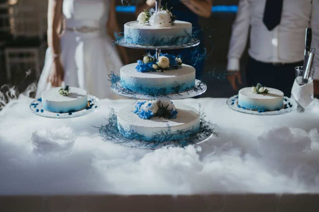 Háromszintes torta asztalon fehér krémmel, kék virágokkal, szárazjéggel körülvéve.