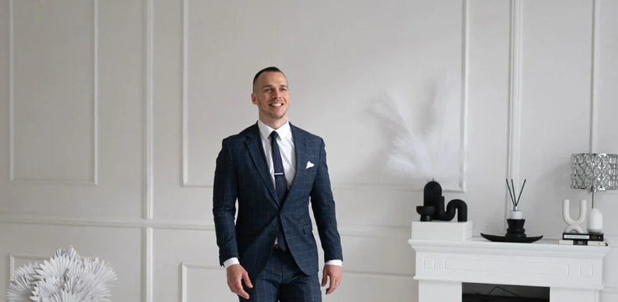 Férfi sötét kockás öltönyben világos minimalista szobában, kandallóval és növényekkel.