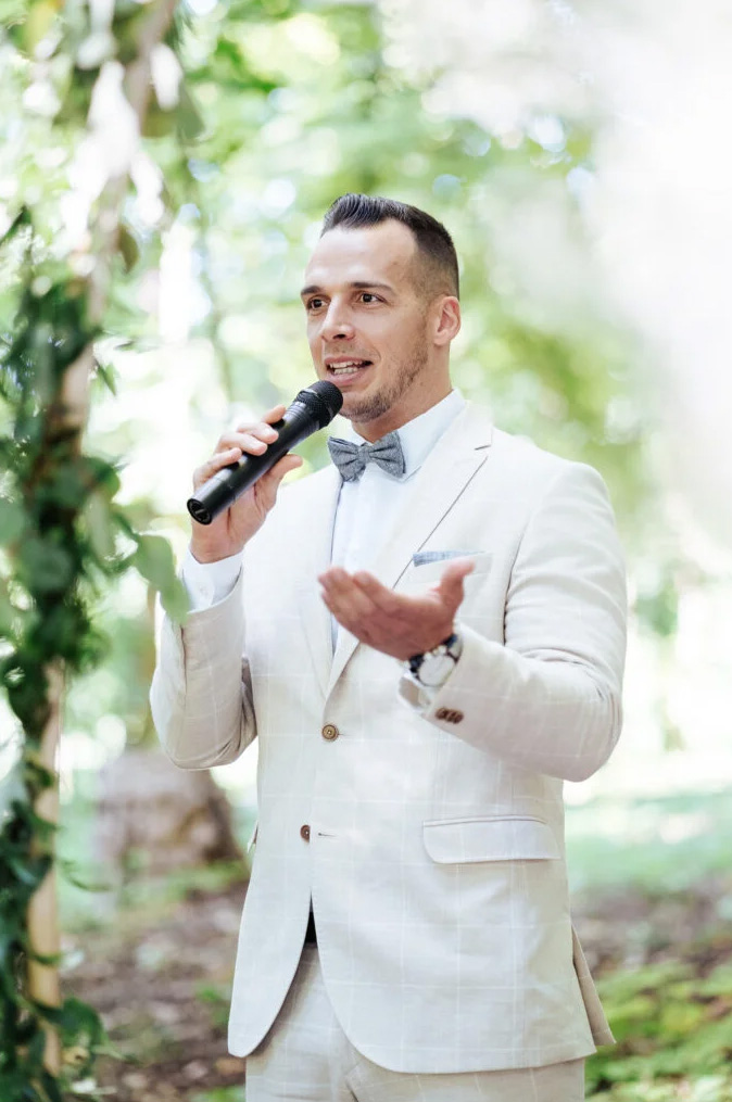 Férfi fehér öltönyben, szürke csokornyakkendővel mikrofont tartva szabadtéri beszédet mond.