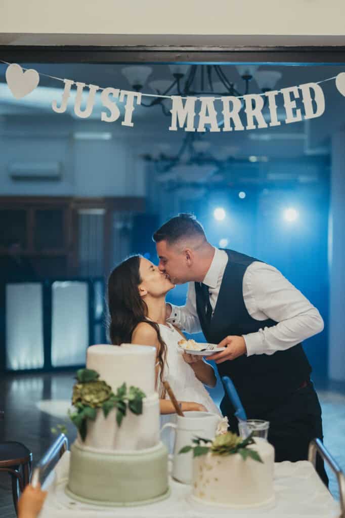 Pár csókolózik esküvői torta előtt, "Just Married" felirat a háttérben.