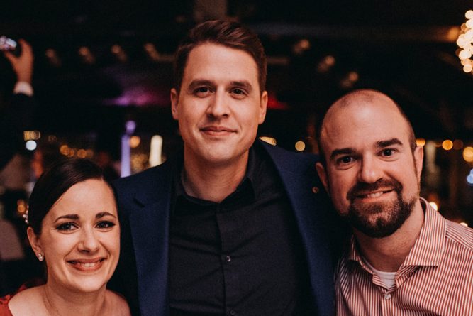 Három felnőtt csoportkép egy beltéri eseményen, háttérben világító dekorációval.