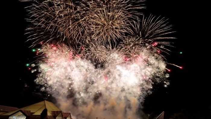 Tűzijáték robban szét az éjszakai égbolton, alul épületek teteje látható.