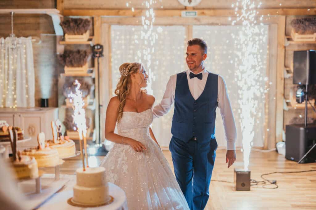 Bride in white dress and groom in navy suit with cakes, fairy lights, and fireworks in background.