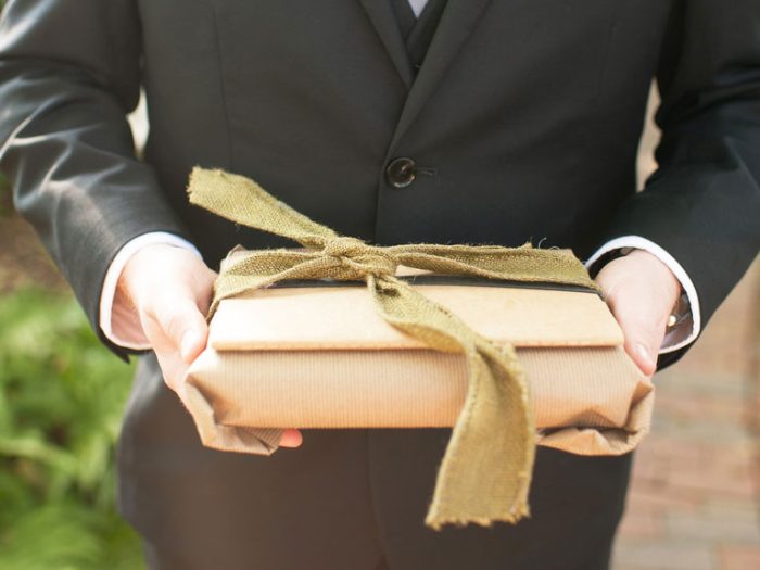 Személy fekete öltönyben barna papírba csomagolt ajándékot tart zöld szalaggal átkötve.