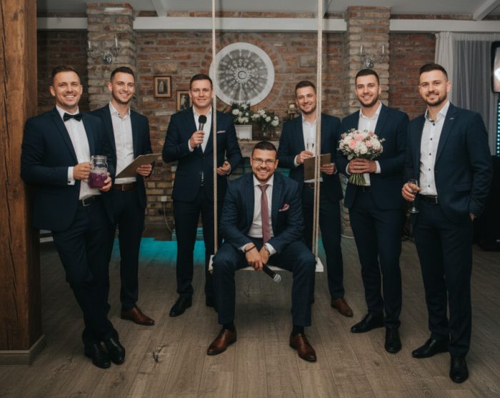 Hét öltönyös férfi beltéri helyiségben, egy hintán ül, két mikrofont, egy virágcsokrot, többen italt tartanak.