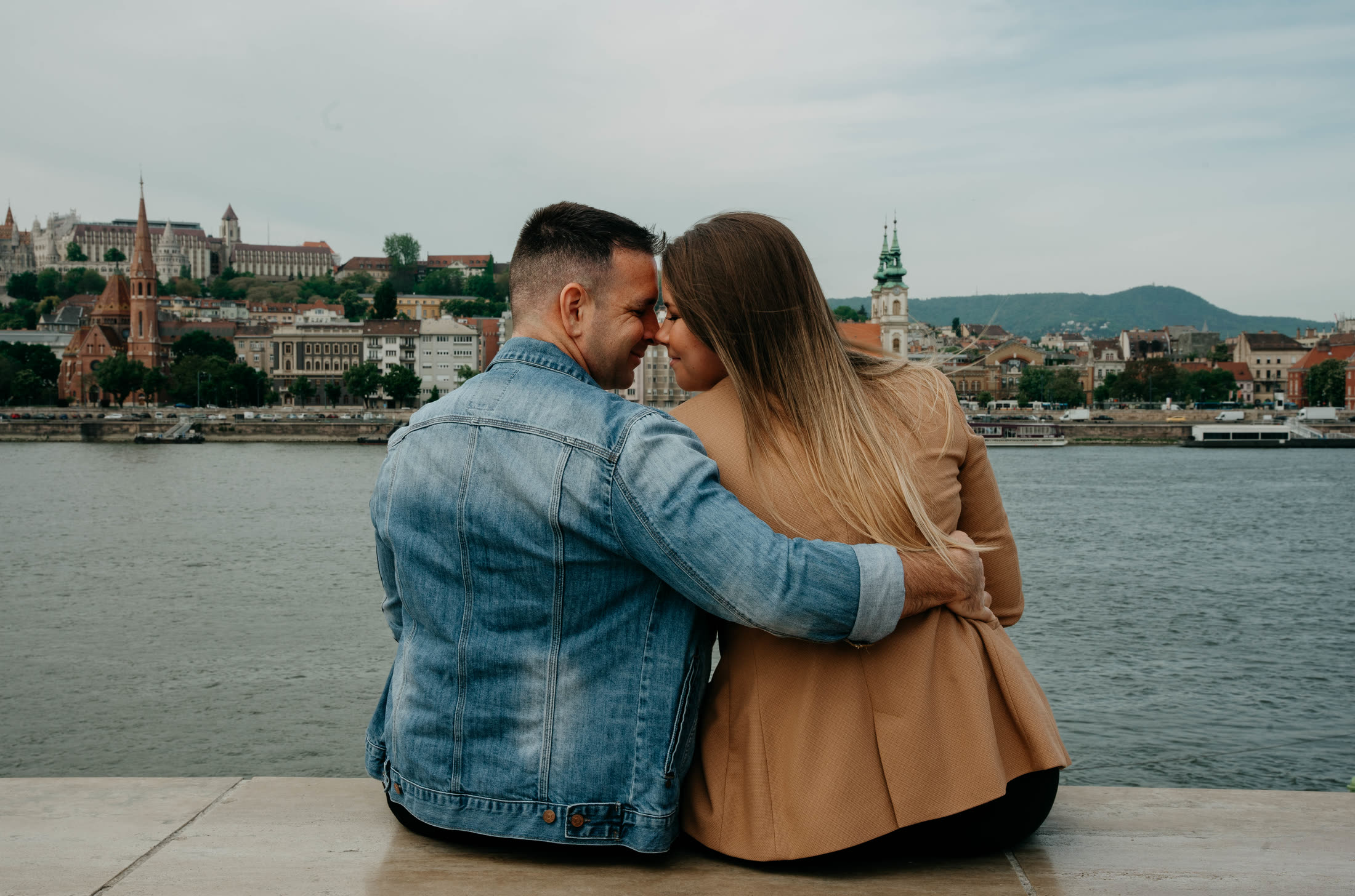 Online esküvői meghívó minta - feree & dóri 1 Feree és Dóri egymást átölelve ül a Duna-parton, háttérben a budai oldal - online esküvői meghívó mintaoldal